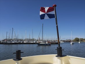 A boat sails in a harbour with a coloured flag on board and many yachts in the background,
