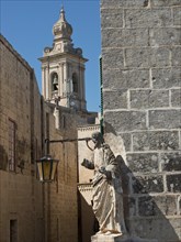 Street with historic architecture, sculpture and church tower in the background, the town of mdina