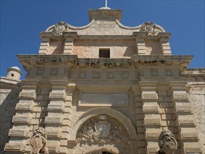 A historic Tor tor with lion sculptures and coats of arms under a blue sky, the town of mdina on
