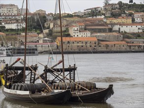 Two wooden boats with barrels in the water, the city and the hilly houses in the background under a