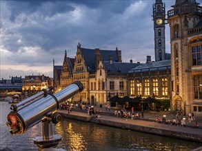 A telescope points to illuminated historic buildings by a river at dusk, blue hour in a medieval