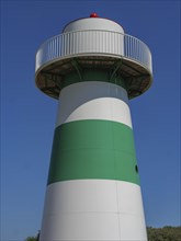 Lighthouse in green and white against a bright blue sky, dunes and beach huts on the beach of