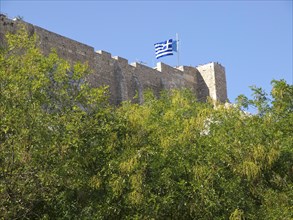 Fortress with Greek flag surrounded by green trees under a clear blue sky, Ancient buildings with