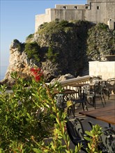 Terrace with chairs and tables next to a rock formation with sea view and historic fortress, the