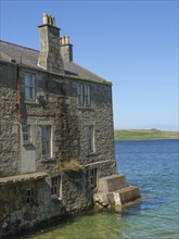 Old stone house on the shore, its foundations partially lapped by clear blue-green water, grey