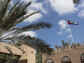 Two flags of the United Arab Emirates waving in the wind, surrounded by palm trees and traditional