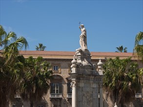 Large religious statue surrounded by palm trees in front of a historic building in sunny weather,
