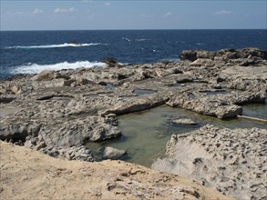 Rugged coastline with rocky pools and the blue sea in the background, the island of Gozo with