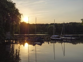 Sailboats at the jetty at sunset, reflected in the water of a calm lake, surrounded by trees, jetty