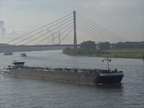 Cargo ship navigating the river under a prominent bridge, industry in the background, shipping