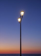 An illuminated street lamp in front of an orange sky at sunset, sunset in many colours on the beach