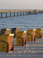 A pier stretches into the sea, surrounded by beach chairs on the sandy beach in the warm evening