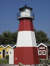 A red and white striped lighthouse in front of a blue sky, surrounded by small colourful wooden