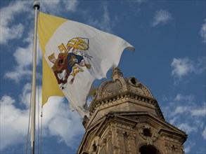 Yellow and white flag with symbols waving in the wind in front of a historic building and blue sky,