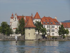 A castle with a red roof on the shore of a lake, a tower and dense trees, sky in the background,