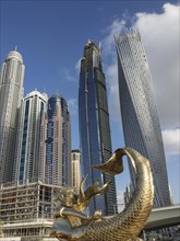 View of modern skyscrapers with a golden mermaid statue in the foreground and a clear blue sky,