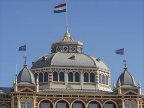 Historic building with Dutch flag and dome, parasols and a pier on the beach of Scheveningen,