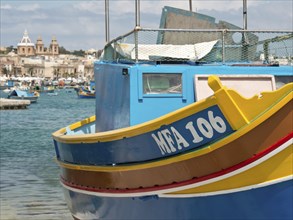 A blue and yellow fishing boat at a harbour with a coastal town in the background, many colourful