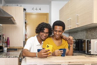 Gay latin adult couple sharing mobile phone while drinking morning coffee at home
