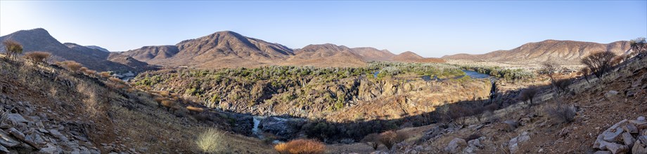 Panorama, Kunene River with green vegetation in dry red mountain landscape, waterfall and African