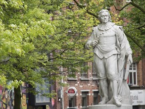 Stone statue of a man in historical clothes in a city park with trees and buildings in the