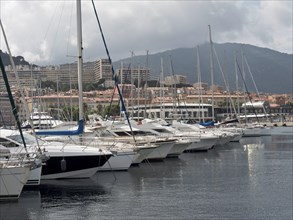 Marina with many parked yachts, behind it a town and mountains under a cloudy sky, Corsica,