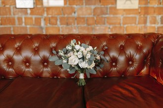 A bouquet of white flowers is placed on a brown leather couch against a brick wall