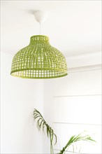 Natural wicker lamp on the ceiling of a white-walled house with a large green plant on the wall
