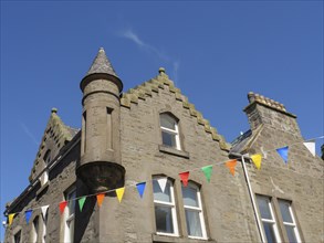 Colourful flags hang around a traditional building with a small tower and windows under a blue sky,
