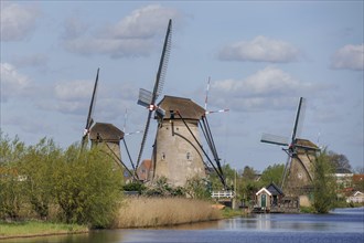 Three windmills next to a canal with trees and village in the background, under a clear sky,