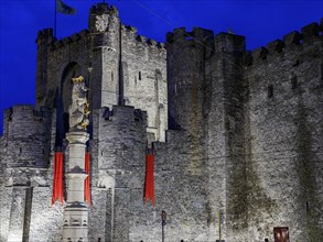 Medieval castle with massive stone walls, a statue and red flags illuminated at night, historic