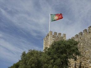 Part of a castle with the Portuguese flag on a watchtower spire, surrounded by green spaces and