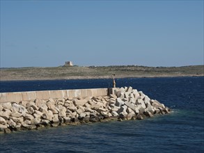 A breakwater of large stones in the sea with a rocky coastline in the background, the island of