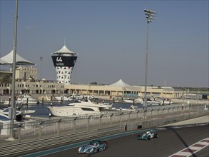 Racing cars drive past a marina, buildings and a striking tower can be seen in the background, Abu