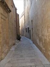 A narrow lane with stone buildings and a cobbled path, quiet and shady, the village of mdina on the