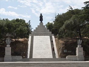 Large monument with a statue on steps, surrounded by trees and a clear sky in the background,