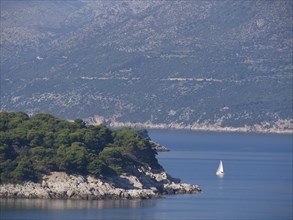 View of a calm sea with a sailing boat and forested mountains in the background, the old town of
