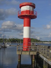 Close-up of a red and white lighthouse on a rough wooden jetty with blue sky and clouds, harbour