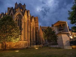 A gothic church with trees and other buildings at dusk, illuminated and under a partly cloudy sky,