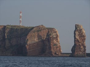 Large red rock cliffs in the sea with a radio tower, accompanied by flying birds against a clear