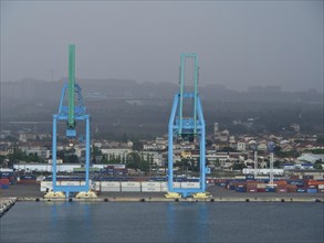 Two large blue harbour cranes in front of a cloudy industrial landscape, several containers in the