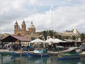 Boats and restaurants in the harbour with church and palm trees in the background on a cloudy day,