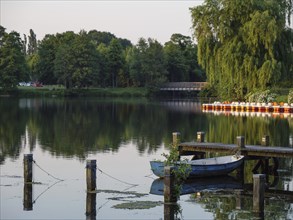 A blue boat is moored at a jetty on calm water, surrounded by green nature, green trees and bushes