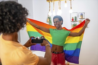 Gay friends taking pictures with camera while waving LGBT flag at home