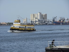 A yellow paddle steamer sailing in the water, harbour facilities and other ships in the background,