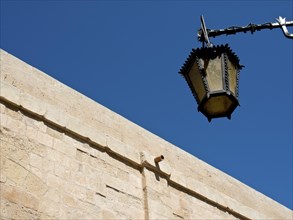 An ancient lantern hangs on a stone wall against a deep blue sky, the town of mdina on the island