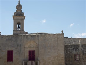 Ancient building with a bell tower and striking red shutters under a clear sky, the town of mdina