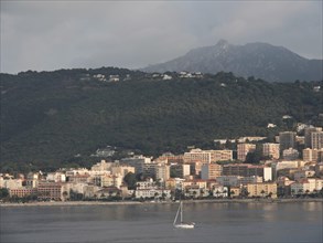 Coastal town at the foot of a green mountain, single boat in the sea under a cloudy sky, Corsica,