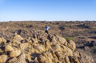 Young man jumping from rock to rock, barren landscape with rocky hills and acacias, African