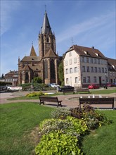 Large Gothic church next to a well-kept town square with benches and flower beds, historic house